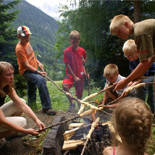 Eine Gruppe von Kindern sitzt um ein Lagerfeuer in der Natur. Sie rösten Stöcke über den Flammen und genießen die Zeit im Freien.