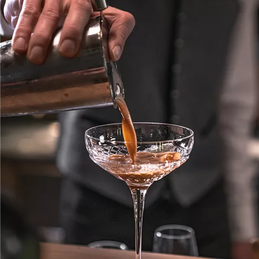 A bartender pours a cocktail into an elegant glass. The background is blurred, which emphasizes the drink and the professional preparation.