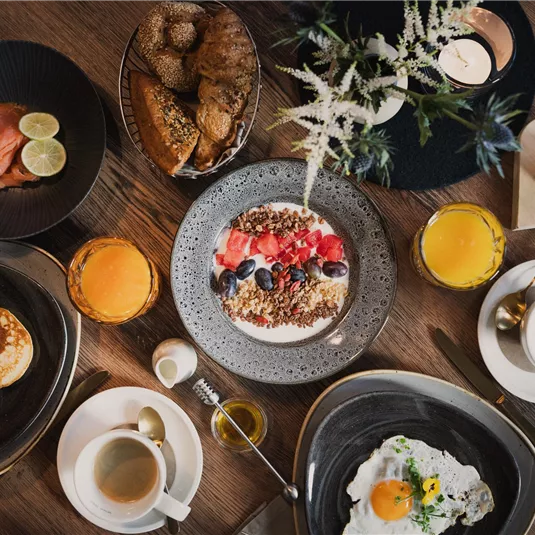 A beautifully set breakfast table with various dishes. There are pancakes, scrambled eggs, fruits, and drinks.