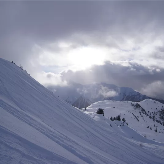 Eine verschneite Berglandschaft mit Wolken am Himmel. Die Sonne scheint schwach durch die Wolken.