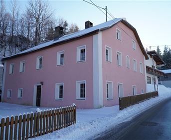 A pink house stands in a snowy landscape. The road runs alongside the building, surrounded by trees.