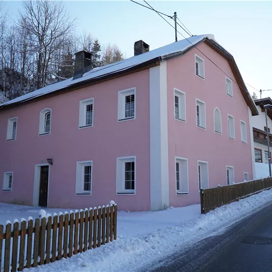 A pink house stands in a snowy landscape. The road runs alongside the building, surrounded by trees.