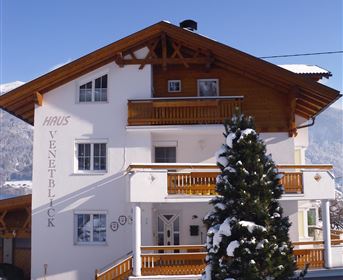 Ein malerisches Holzhaus im Schnee, umgeben von einer verschneiten Landschaft. Die Fassade ist hell und freundlich mit einem kleinen Balkon und einem grünen Baum im Vordergrund.