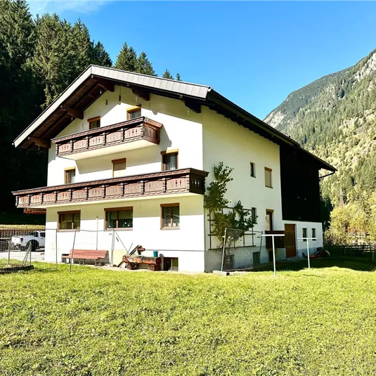 A large house in the mountains, surrounded by trees and green meadows. The architecture is traditional with balconies and a clear blue sky in the background.