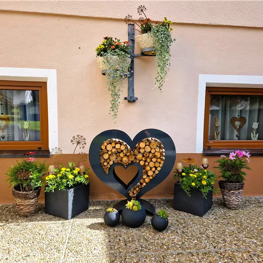 A cozy corner of the house with two windows and colorful flowers in pots. In the foreground is a large heart made of wooden slices.