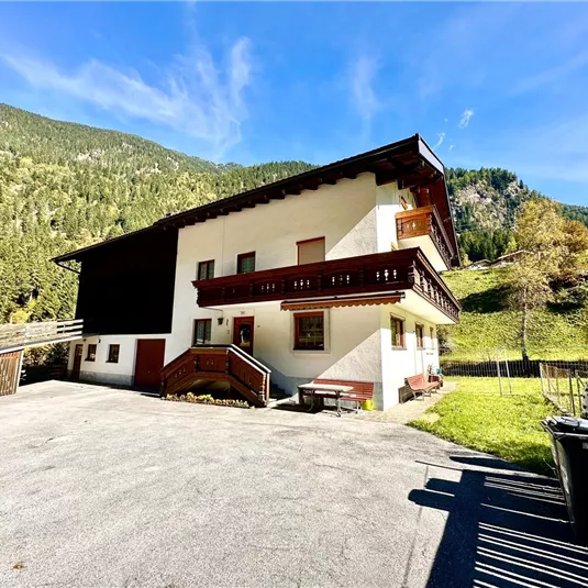 A charming house in the mountains with a spacious parking area. Surrounded by green forests and a clear blue sky backdrop.