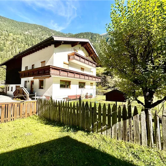 A cozy house with a balcony and triple windows, surrounded by green meadows and mountains. A well-maintained garden with a wooden picket fence completes the rural atmosphere.
