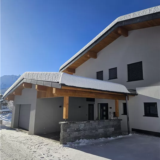 A modern house with a wooden roof and a stone wall in the foreground. The sky is clear and blue, and snow is on the ground.