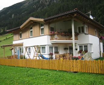 Ein schönes Haus im alpinen Stil mit einem Garten und Holzzäunen. Die Umgebung ist grün mit Bergen im Hintergrund.