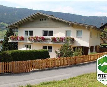 A beautiful residential house with flowers on the balcony, surrounded by green scenery. In the background, mountains can be seen.