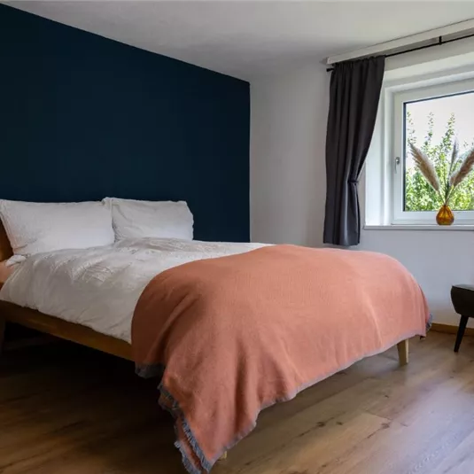 Ein modernes Schlafzimmer mit einem gemütlichen Bett und orangefarbener Decke. Der Raum hat ein Fenster mit Blick auf die Natur und einen eleganten Sessel.