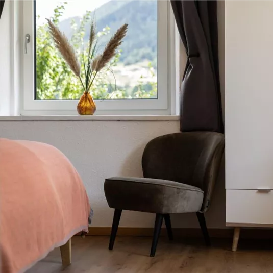 Ein gemütliches Zimmer mit einem rosa Bett und einem schwarzen Sessel. Der Blick aus dem Fenster zeigt eine grüne Landschaft und Pflanzen.