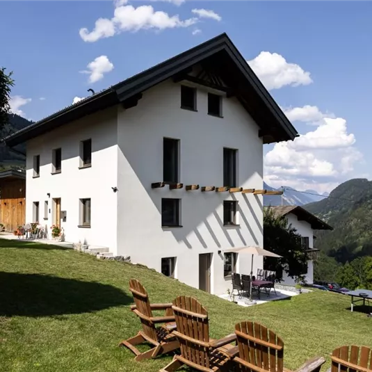 Ein modernes Haus mit einem schönen Garten in den Bergen. Im Vordergrund stehen Holzstühle und der Blick auf die Landschaft ist atemberaubend.