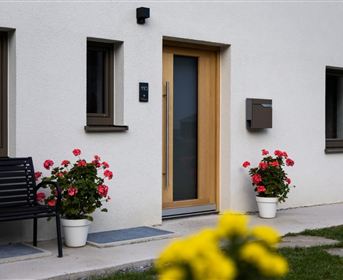 A modern house with a wooden front door and large windows. On the terrace, blooming flowers in pots are standing, and a bench invites you to sit down.
