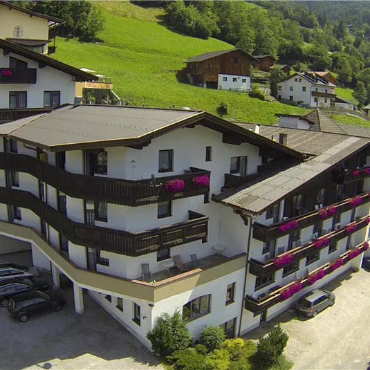 A modern hotel in a green landscape with mountains in the background. The facade has balconies with blooming flowers and there are parking spaces in front of the building.
