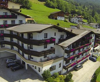 Ein modernes Hotel in einer grünen Landschaft mit Bergen im Hintergrund. Die Fassade hat Balkone mit blühenden Blumen und es gibt Parkplätze vor dem Gebäude.