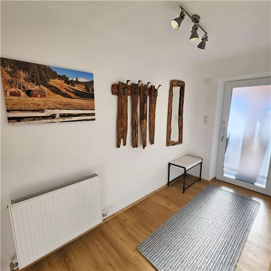 A friendly entrance area with wooden flooring and a gray doormat. Rustic coat hooks and a wall mirror are hanging on the wall, while a landscape picture decorates the wall.