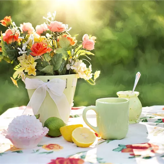 A beautiful table with a flower vase, lemon-yellow citrus fruits, and a cup. In the background, the sun is shining and the greenery creates a fresh atmosphere.