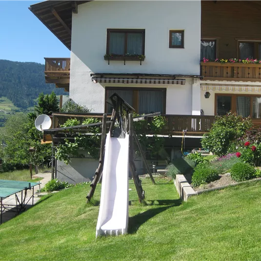 A beautiful house with a garden and a slide. In the background, green hills can be seen.