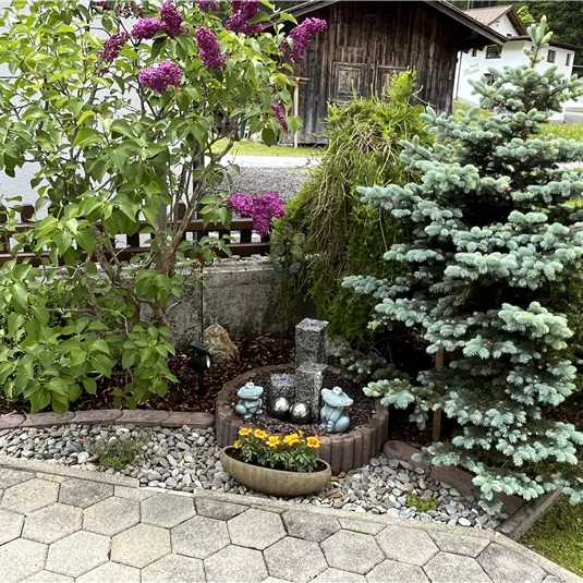 A beautiful garden area with purple lilac and a blue conifer. In the middle, there is a small fountain with colorful flowers.