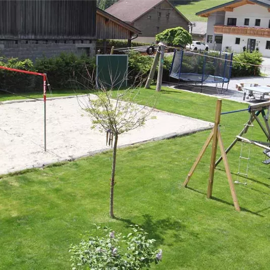 Ein gepflegter Garten mit einem Sandvolleyballfeld und einem Spielplatz. Im Hintergrund sind Wohnhäuser und eine grüne Landschaft zu sehen.