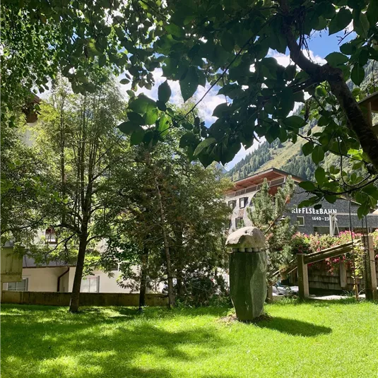 A green meadow with trees in the foreground and a building in the background. The sky is blue with some clouds.