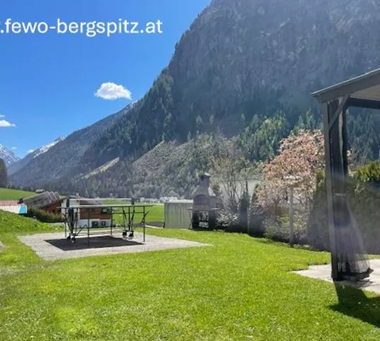 A green meadow with a view of majestic mountains and blue sky. In the foreground, there is a terrace with a grill.
