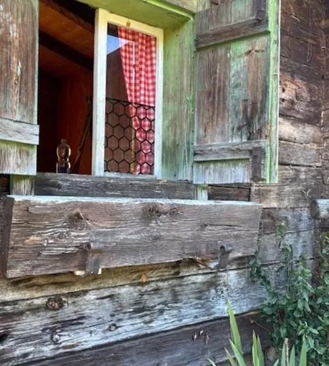 Ein altes Holzhäuschen mit einem offenen Fenster. Die Fensterläden sind grün, und im Inneren sind rote Karos angebracht.