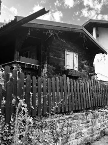 Ein traditionelles Holzhaus mit einem Gartenzaun in einer malerischen Umgebung. Der Himmel ist teilweise bewölkt und die Landschaft wirkt ruhig.