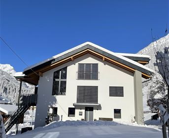 Ein modernes Haus im Schnee mit einem klaren, blauen Himmel. Die Umgebung ist von Bergen umgeben, und der Weg ist schneebedeckt.