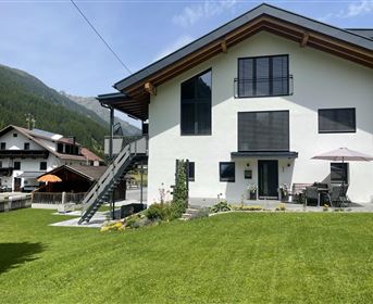 Ein modernes Haus mit einem gepflegten Garten und einer Terrasse. Im Hintergrund sind Berge und weitere Gebäude sichtbar.