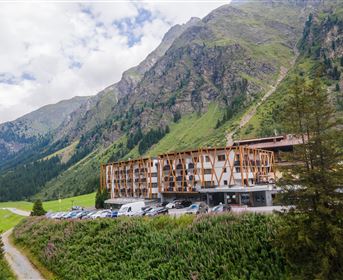 Ein modernes Hotel inmitten einer beeindruckenden Berglandschaft. Grüne Wiesen und mächtige Berge umgeben die Unterkunft.
