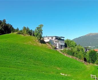 Ein modernes Haus auf einer grünen Wiese umgeben von Bäumen. Im Hintergrund sind Berge und ein klarer blauer Himmel sichtbar.