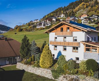 Ein modernes Haus in einer malerischen Almlandschaft mit schönen Gärten. Im Hintergrund sind grüne Hügel und traditionelle Gebäude zu sehen.