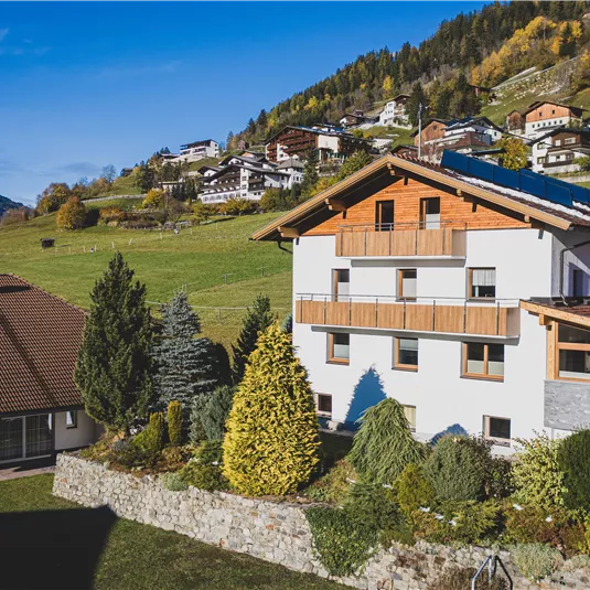 Ein modernes Haus in einer malerischen Almlandschaft mit schönen Gärten. Im Hintergrund sind grüne Hügel und traditionelle Gebäude zu sehen.
