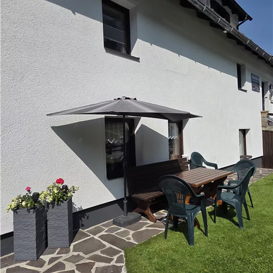 Ein schöner Garten mit einer Terrasse und einem Sonnenschirm. Am Tisch stehen Stühle und blühende Pflanzen in Töpfen.