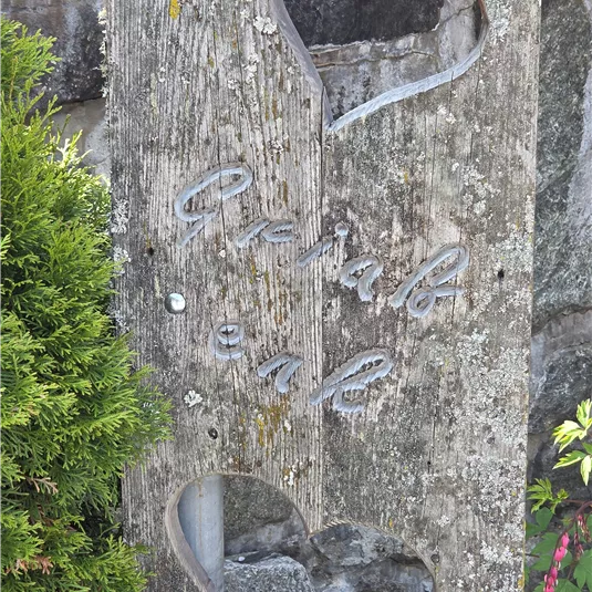 Ein Holzschild mit herzförmigen Ausschnitten und der Aufschrift „Er-Häb eal“. Es steht in einer grünen Umgebung mit Pflanzen.