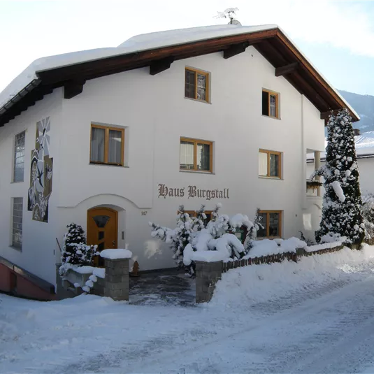 A charming, snow-covered house with a white facade. The path is gently curved and lined with snow-covered trees.