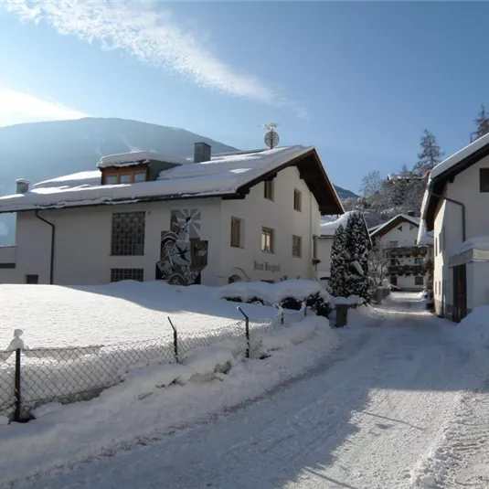 A snow-covered landscape with several houses and a quiet path. In the background, mountains and a blue sky are visible.