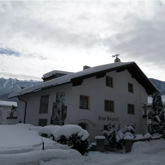 A beautiful, snow-covered house in a winter landscape. In the background, there are mountains and a cloudy sky.