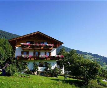 A beautiful, traditional house in the mountains. It is surrounded by green meadows and trees, with blooming flower boxes at the windows.