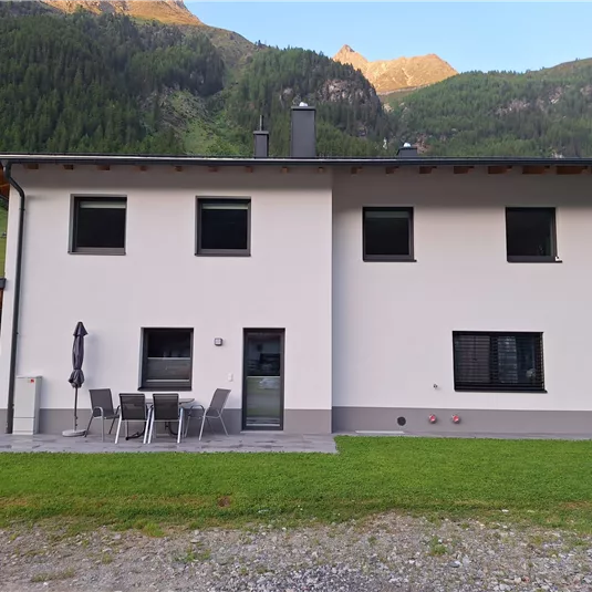 Ein modernes zweigeschossiges Haus mit einer hellen Fassade und großen Fenstern. Im Vordergrund steht eine Sitzgruppe und der Hintergrund zeigt eine grüne Berglandschaft.