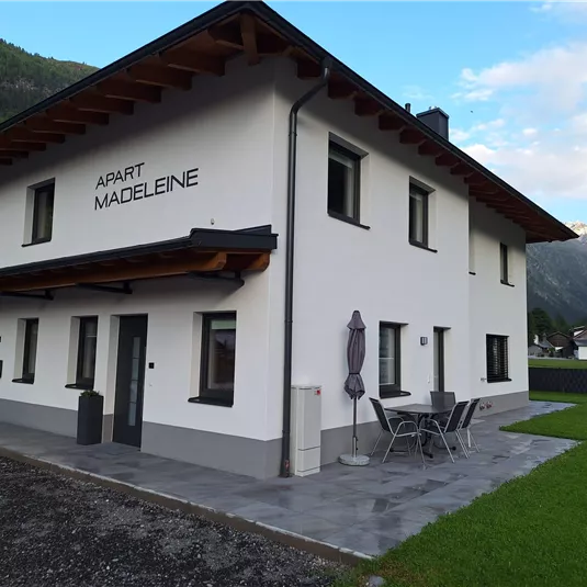 Ein modernes, zweigeschossiges Gebäude mit dem Namen "Apart Madeleine". Vor dem Haus gibt es einen kleinen Garten und eine Sitzgelegenheit.