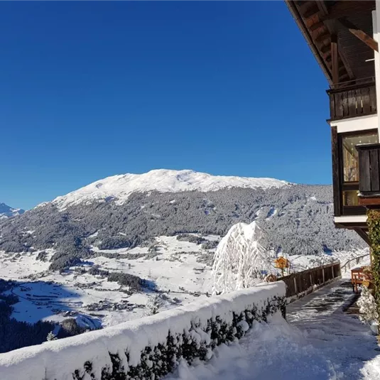 A beautiful winter view of snow-covered mountains and valleys. In the foreground, a traditional wooden house with snow on the balcony.