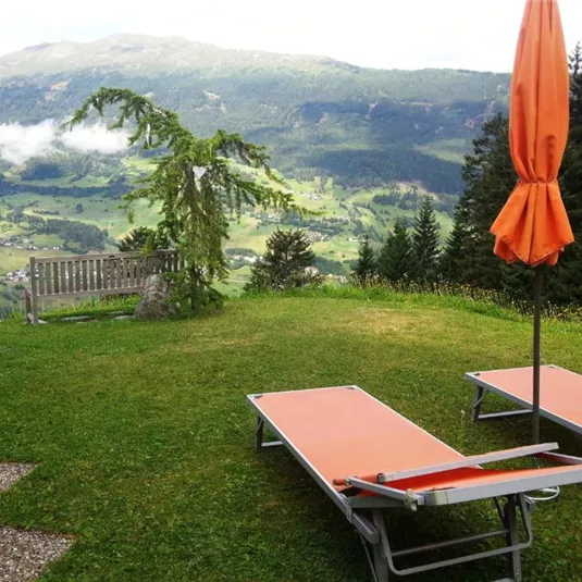 A relaxing view of a green hilly landscape with a sun lounger and a sun umbrella. In the background, mountains and clouds are visible.