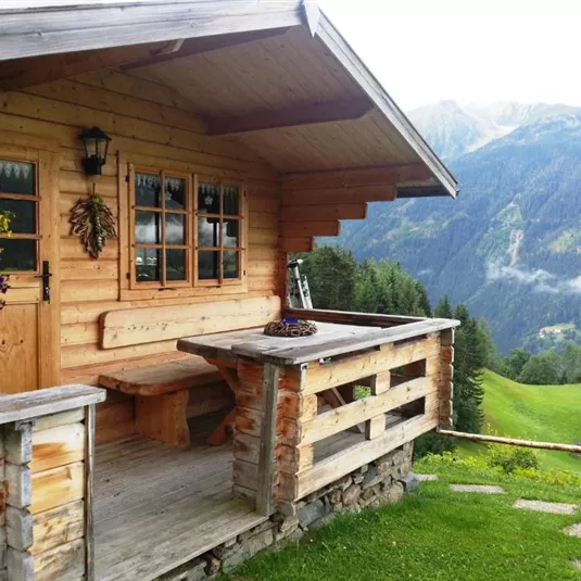 A cozy wooden house with a balcony and beautiful flowers. In the background, green mountains and a clear sky can be seen.