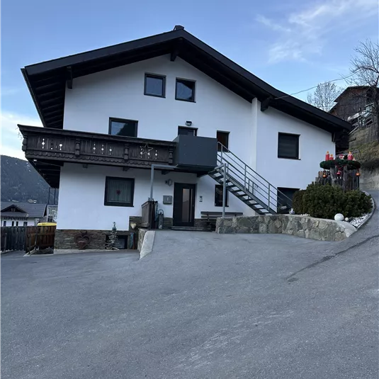 A modern house with a white facade and a balcony. It is located on a small street and is surrounded by a picturesque landscape.