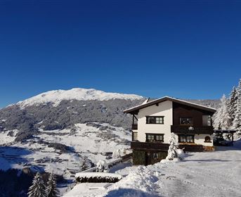 A snow-covered mountainous landscape with a cozy house. In the background, majestic mountains and a clear blue sky can be seen.