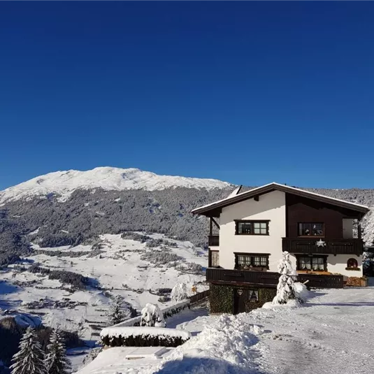 A snow-covered mountainous landscape with a cozy house. In the background, majestic mountains and a clear blue sky can be seen.