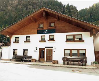 A traditional Alpine house with a wooden facade and several windows. It stands in a picturesque setting with mountains in the background.